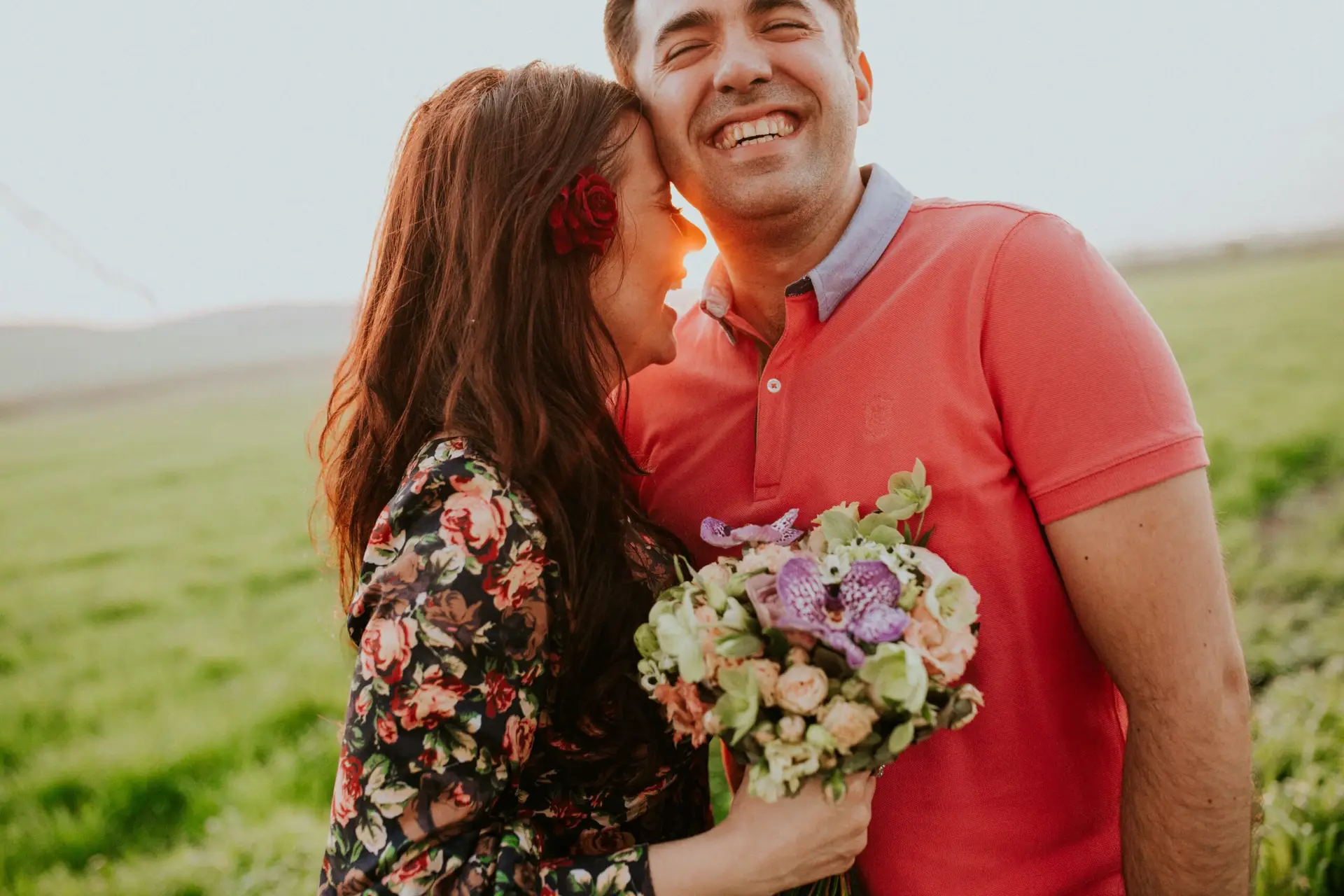 Smiling couple embracing in a sunlit grassy field, woman holding a bouquet of flowers.