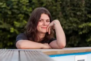 Smiling woman with brown hair resting her chin on her hand outdoors near greenery in early recovery.