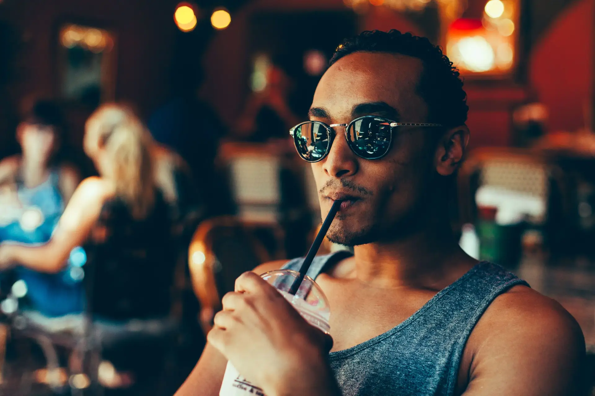 Young man in sunglasses and gray tank top sipping a drink through a straw at a café.