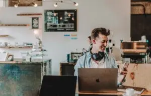 Smiling man with headphones works on a laptop and checks his phone at a coffee shop counter.