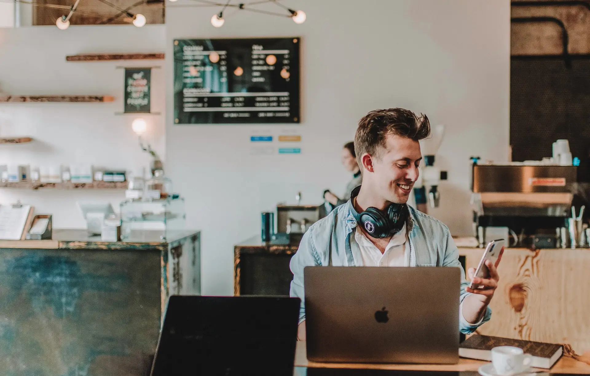 Smiling man with headphones works on a laptop and checks his phone at a coffee shop counter.