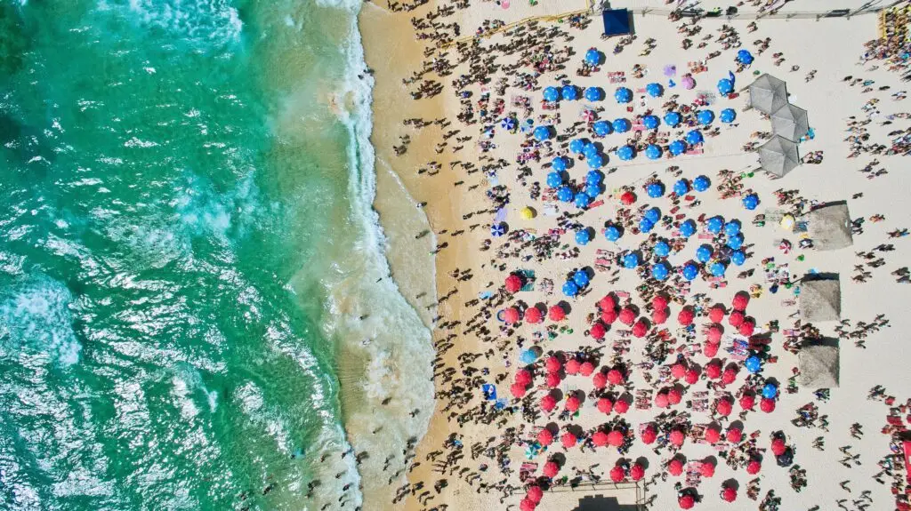 Aerial view of a crowded Fort Lauderdale beach with blue and red umbrellas beside turquoise ocean waves.