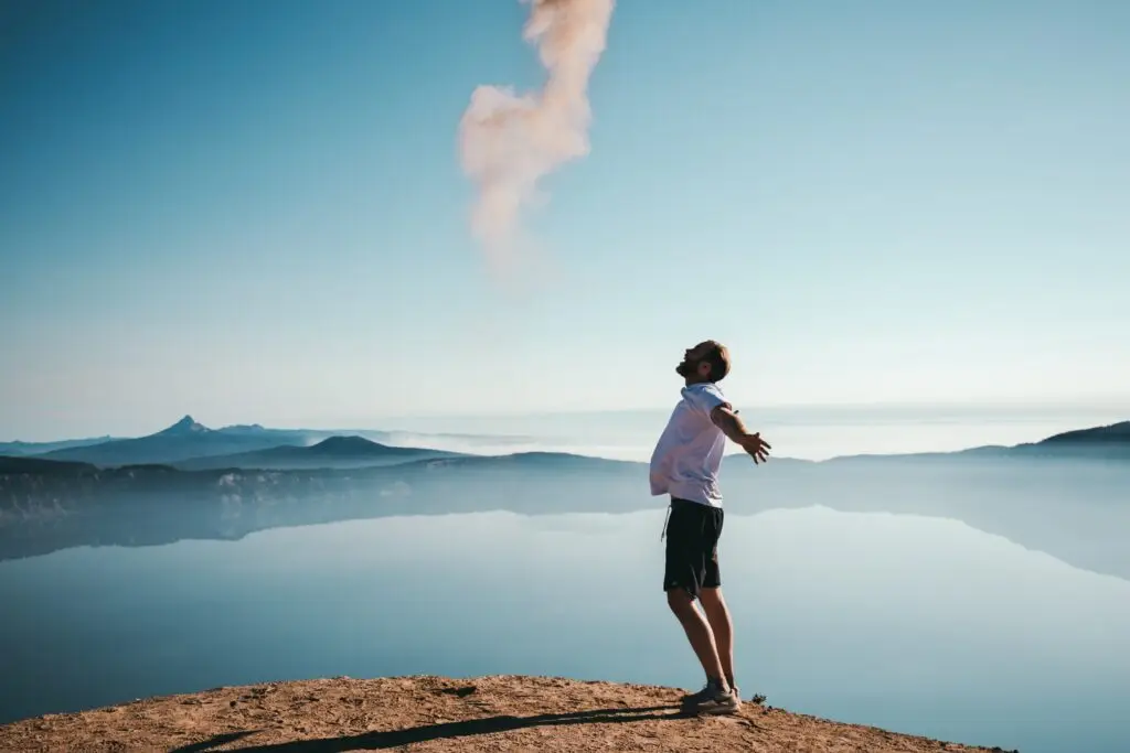 Man standing on a cliff with arms open wide, facing a calm lake and misty mountains under a clear sky.