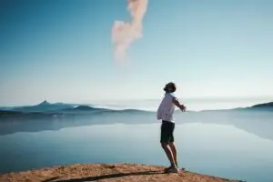 Man standing on a cliff with arms open wide, facing a calm lake and misty mountains under a clear sky.