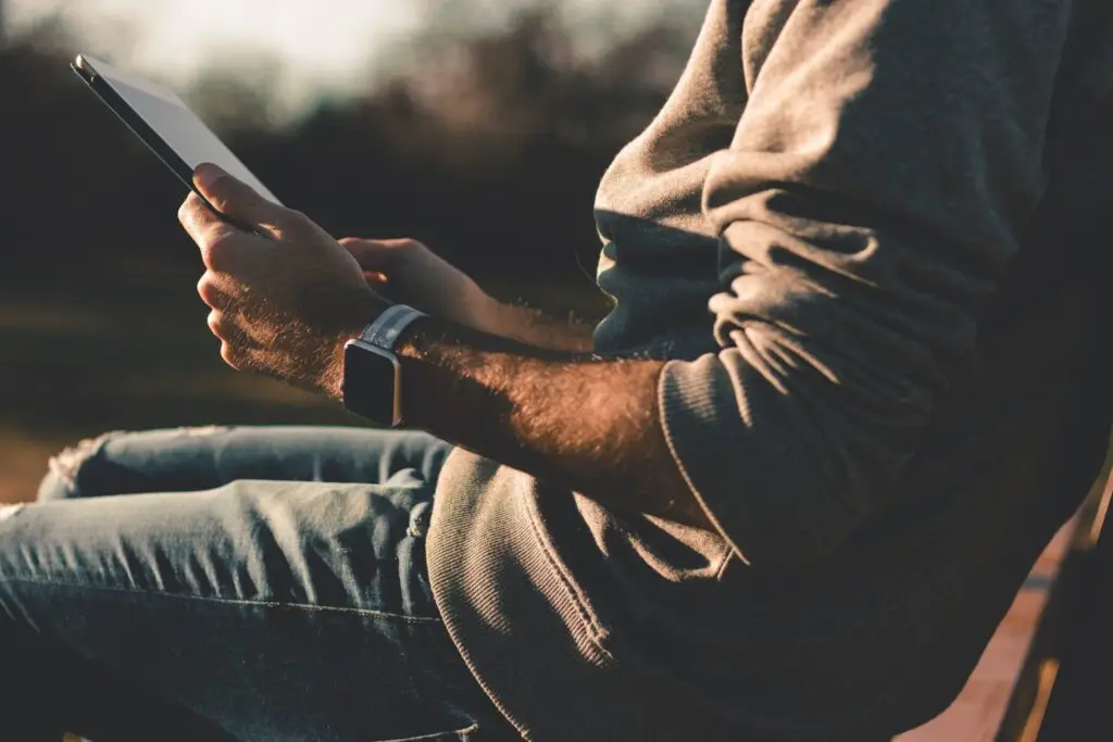 Man sitting outdoors at sunset holding a tablet, wearing a smartwatch and casual clothing.