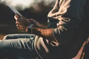 Man sitting outdoors at sunset holding a tablet, wearing a smartwatch and casual clothing.