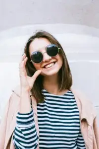 Smiling woman in round sunglasses and striped shirt standing outdoors against a light wall.